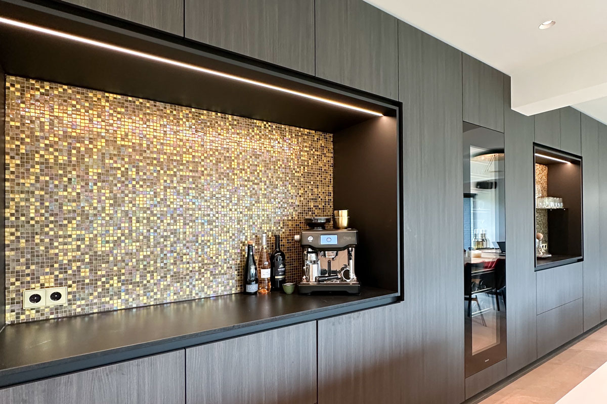 Luxe wandkast in keuken met donkere houten afwerking, geïntegreerde LED-verlichting en gouden mozaïektegels als achterwand. De kast bevat een ingebouwde koffiemachine en stijlvolle opbergmogelijkheden.