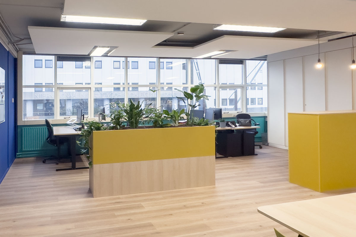 Kantoorinrichting bij Staij in Amsterdam met blauwe accentmuur, gele plantenbak en zit-sta bureaus