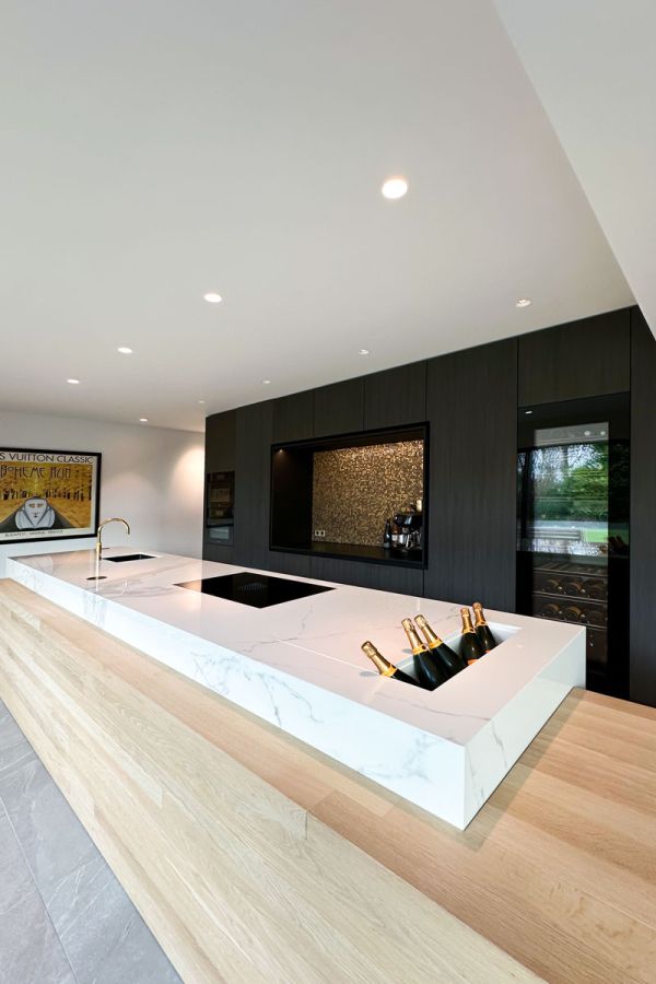 Luxe keuken met een marmeren kookeiland, geïntegreerde champagnekoeler en houten tafelblad. De achterwand is voorzien van gouden mozaïektegels en donkere kastafwerking. Een elegante en functionele combinatie van materialen.