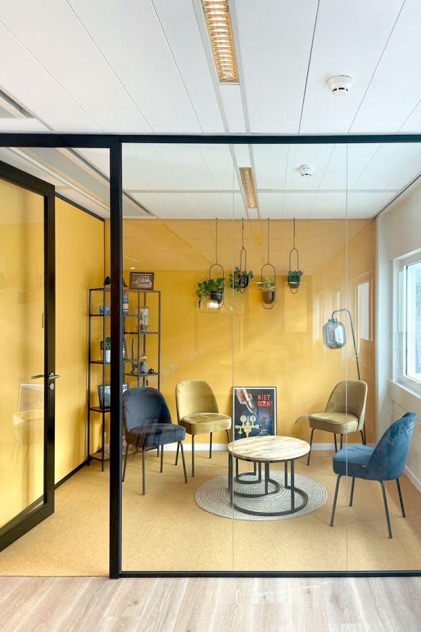 Compacte loungehoek met glazen wanden, gele muur, fluwelen stoelen en een moderne zwarte boekenkast.