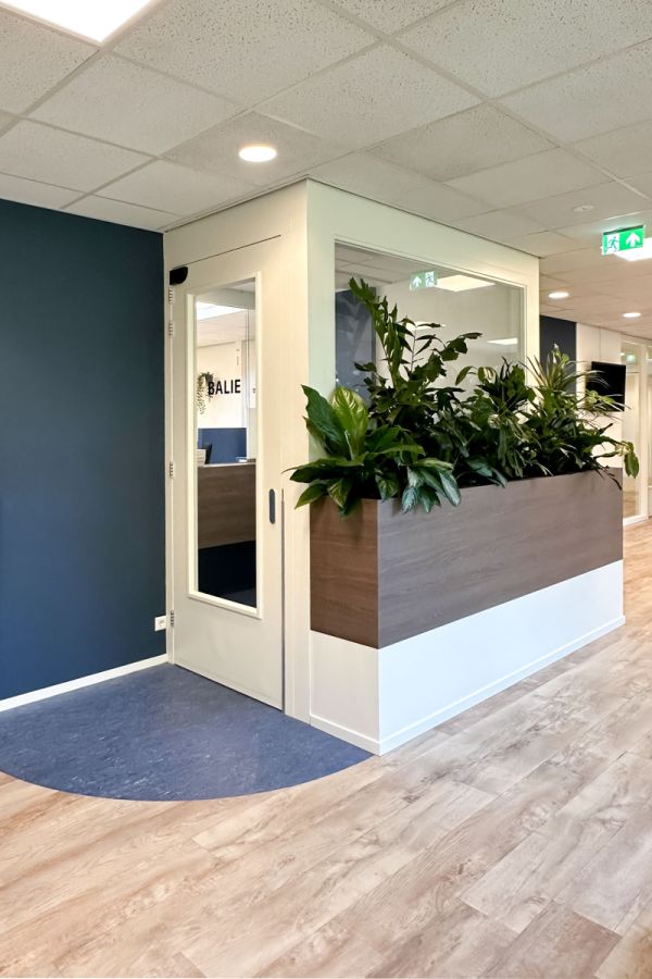 Moderne wachtkamerinrichting met ombouw rondom de balie, geïntegreerde plantenbak en houten look PVC vloer voor een warme sfeer.