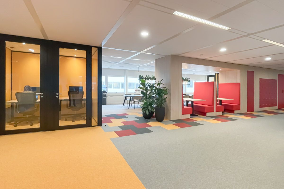 Interieur van een modern kantoor met glazen stilteruimtes, op maat gemaakte rode treinbanken en kleurrijke vloerbedekking met gele, rode en grijze tinten.