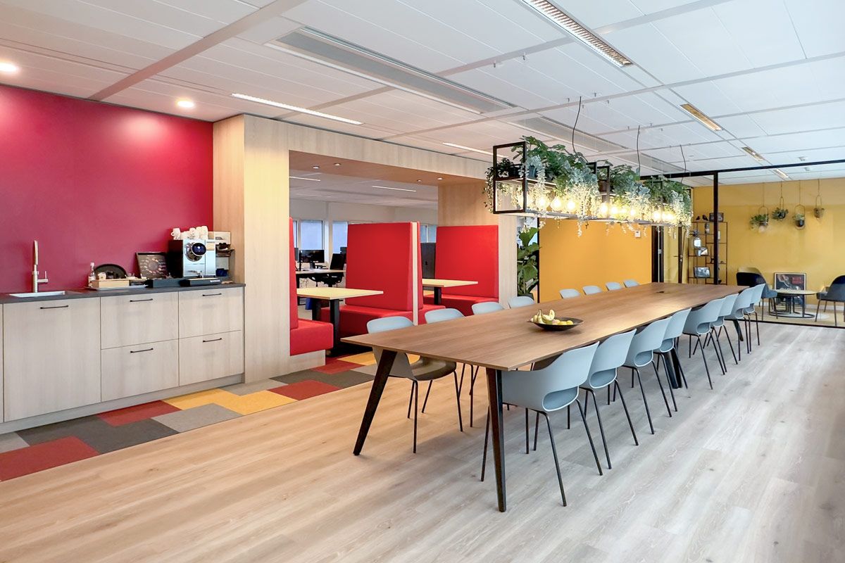 Lunchruimte in kantoor met lange houten tafel, designstoelen, een pantry met koffiemachine en ingebouwde rode treinbankjes. Hanglampen met planten zorgen voor sfeer.