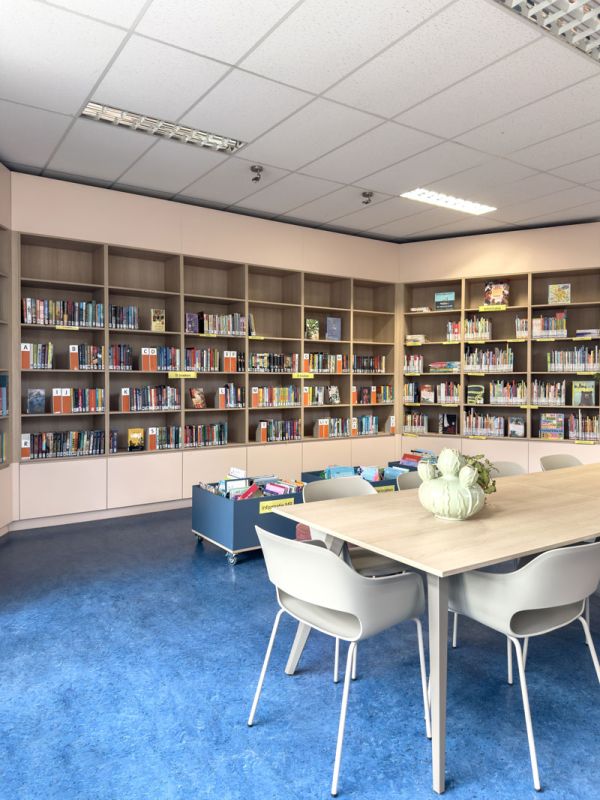 Bibliotheekkast in roze en hout met blauw marmoleum en tafel met stoelen.