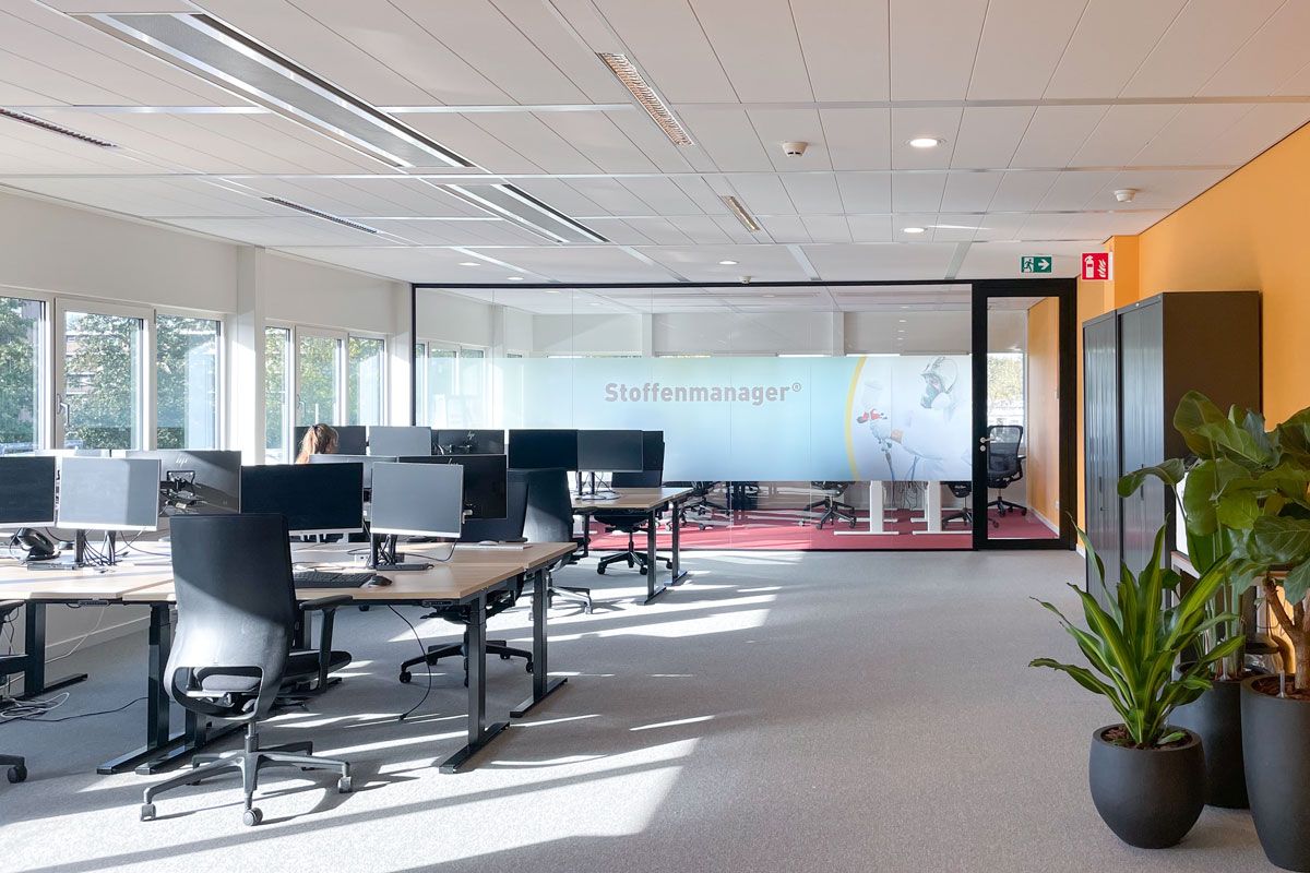 Open kantoorruimte met bureaus, zwart-witte bureaustoelen, en grote ramen die zorgen voor veel daglicht. Achtergrond met glaswand en Stoffenmanager-logo.