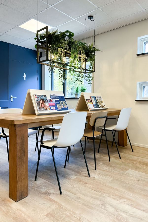 Leestafel in wachtkamer met houten look, tijdschriften en hangende planten. Modern en rustig interieur met veel lichtinval.