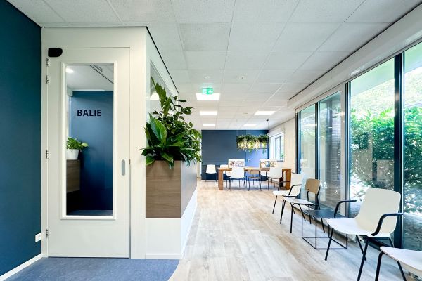 Leestafel in wachtkamer met houten look, tijdschriften en hangende planten. Modern en rustig interieur met veel lichtinval.