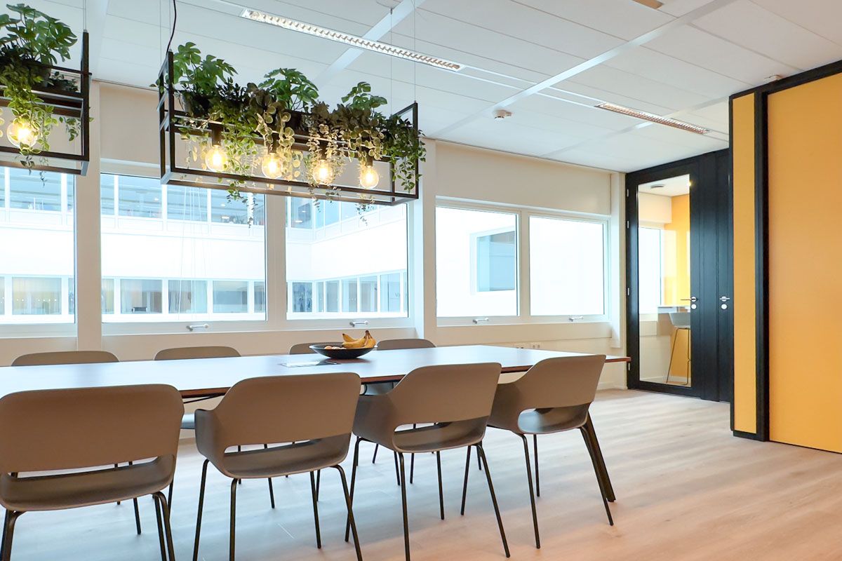 Interieur van een kantoor met een ruime lunchruimte, lange houten tafel, hangende plantenlampen en pantry met rode en gele accenten.