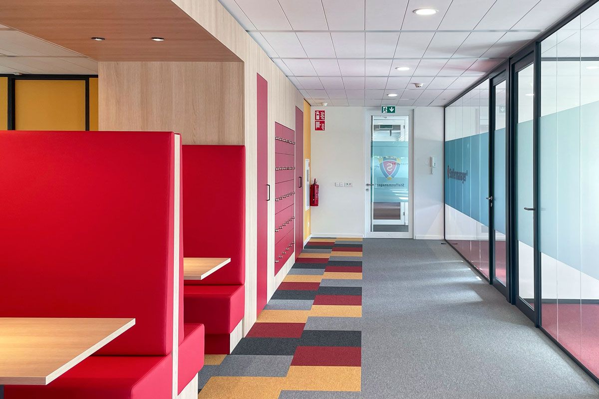 Hal in een kantoor met glazen wanden, rode treinbanken, een kast met kluisjes en een kleurrijke vloerbedekking in rode, gele en grijze tinten.