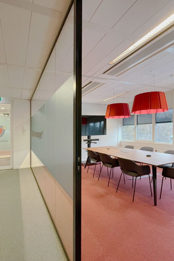 Vergaderruimte met glazen wand, houten tafel, rode hanglampen en rode vloerbedekking.