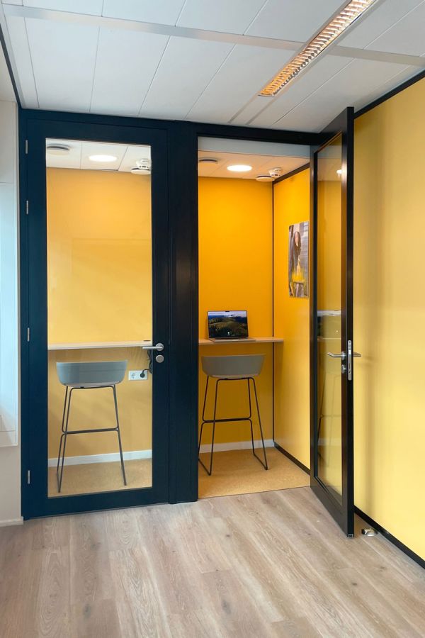 Stilteruimte met glazen deuren, ingericht met barkrukken en werkbladen tegen een gele muur, voorzien van verlichting.