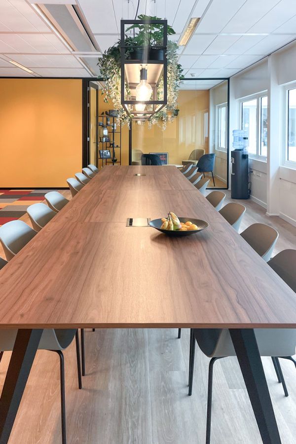 Vergadertafel met houten blad, stoelen en hangende plantenlamp in kantoor met glazen wanden en gele accenten.