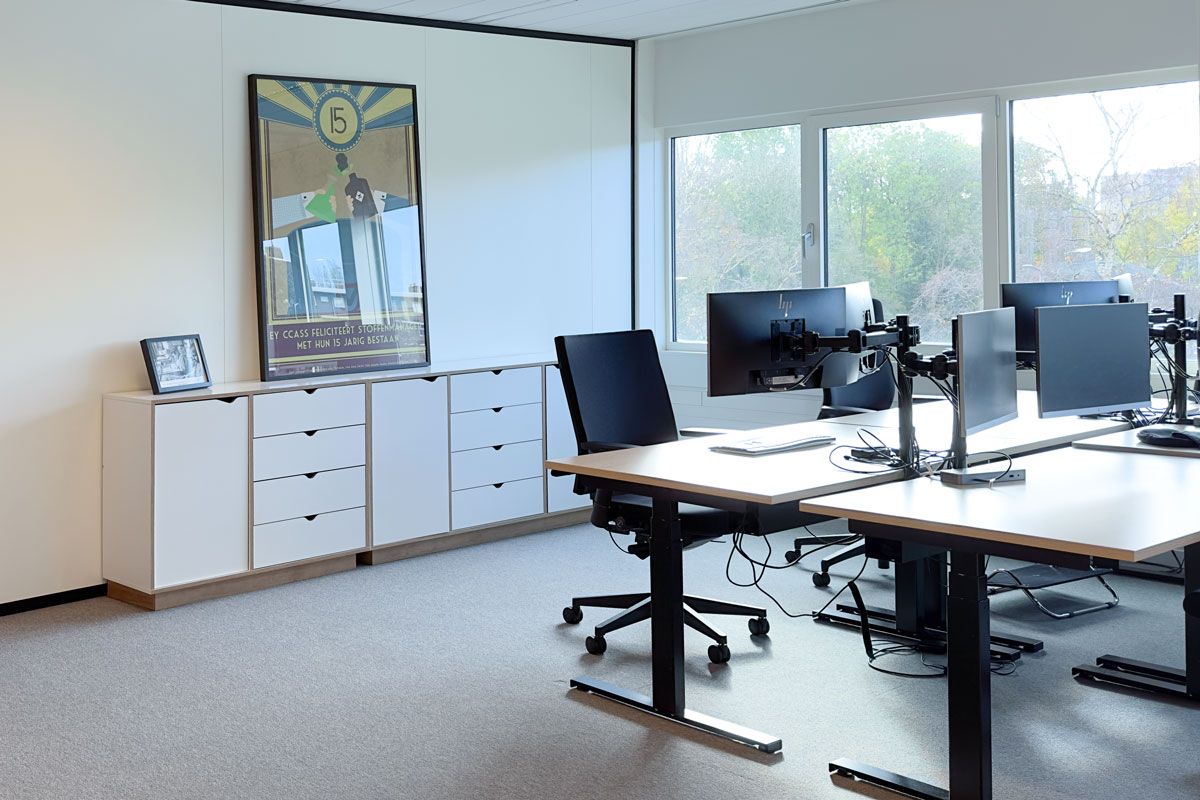 Moderne kantoorruimte met ergonomische bureaus, verstelbare monitorarmen en een opbergmeubel met lades en kasten tegen de muur.