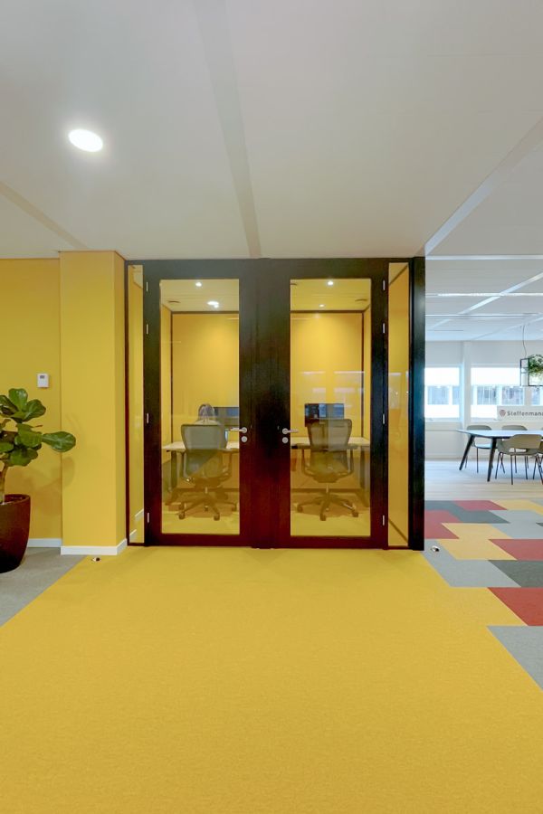 Stilteruimte met glazen deuren en gele muren, ingericht met ergonomische bureaustoelen en werkplekken.
