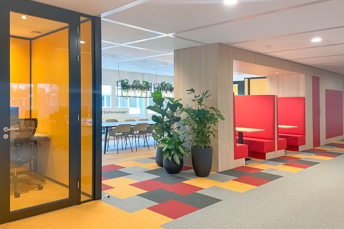 Moderne kantoorruimte met een glazen stilteruimte, een lunchruimte met planten en verlichting, rode treinbanken en een kleurrijke vloer.