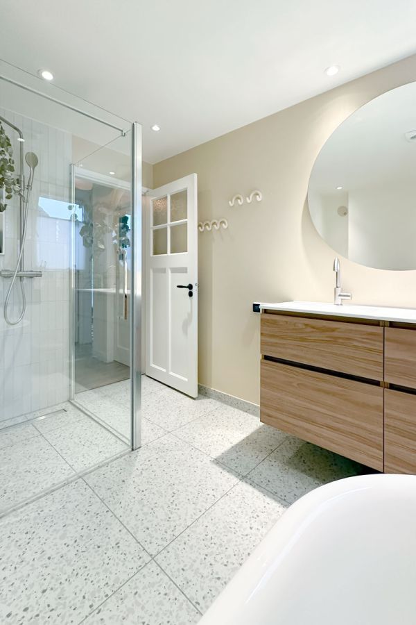 Vergrote badkamer in jaren 30 woning met terrazzo vloer, glazen douchewand en houten wastafelmeubel.