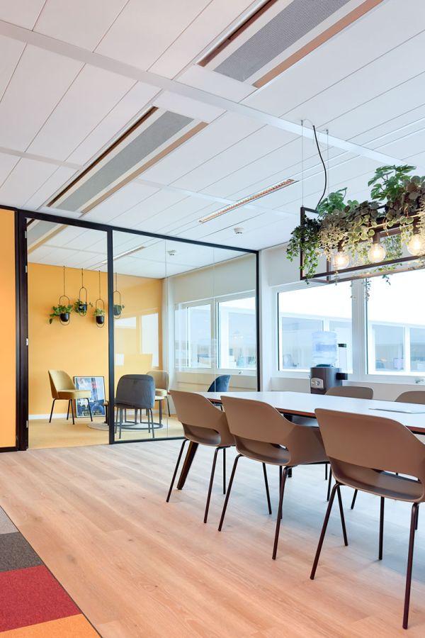 Moderne lunchruimte met glazen wanden, planten en een lange tafel met comfortabele stoelen, naast een vergaderruimte.