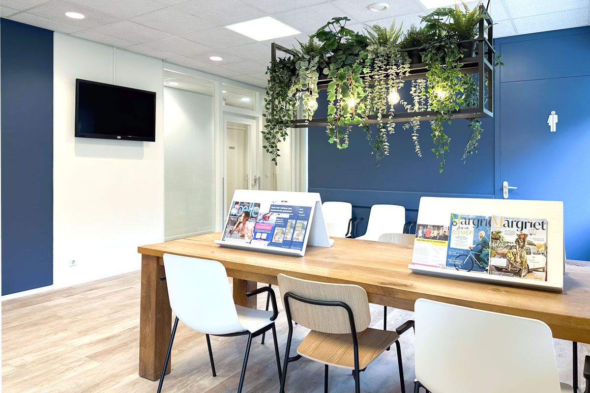 Leestafel met tijdschriften in moderne wachtkamer, houten look PVC vloer en hangende planten voor een frisse en uitnodigende sfeer.