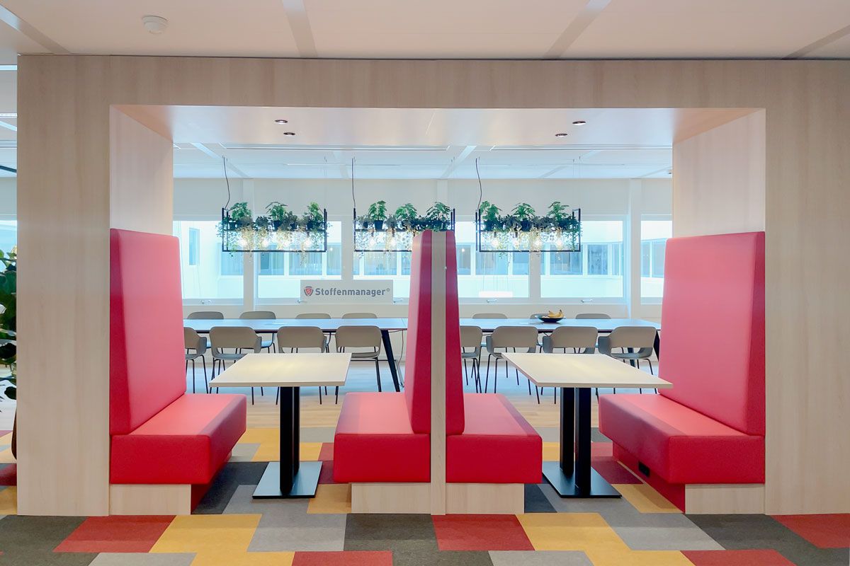 Treinbanken met rode bekleding in een kantoorruimte, met zicht op een vergadertafel en hangende plantenlampen op de achtergrond.