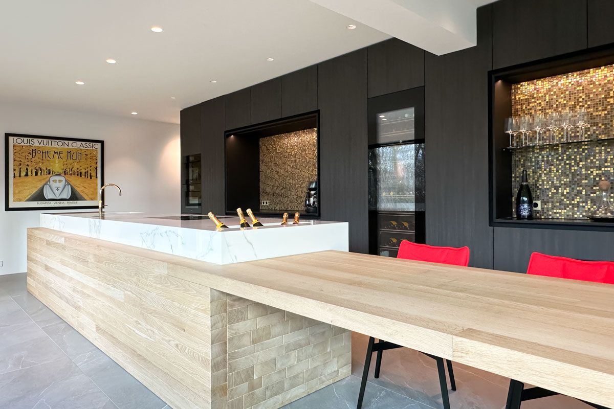Luxe keuken met marmeren kookeiland, eikenhouten tafel, rode stoelen, en gouden details. Inclusief wandkasten met mozaïektegels en glazen wijnrekken.