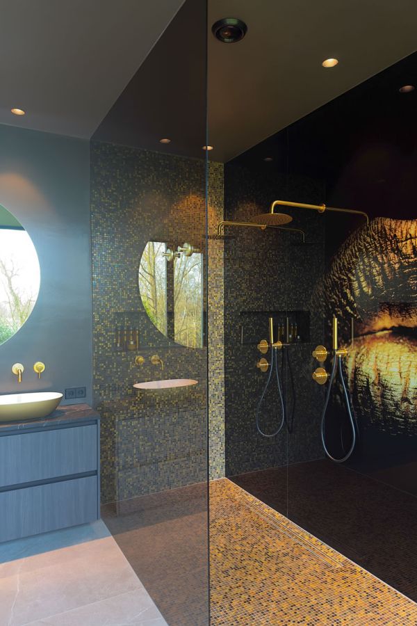 Luxe badkamer met gouden mozaïektegels, dubbele douchekoppen met gouden afwerking, en een opvallende kunstzinnige wandprint. Modern wastafelmeubel met ronde spiegel.