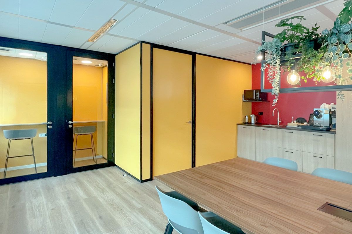 Vergaderruimte met houten tafel, stoelen, glazen wanden, gele muren en keukenblok met hangende plantenlampen.